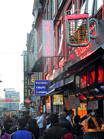 Fotot f&ouml;rest&auml;ller en gatubild fr&aring;n en stadsk&auml;rna. Man ser neonskyltar fr&aring;n restauranger och v&auml;xlingskontor.
